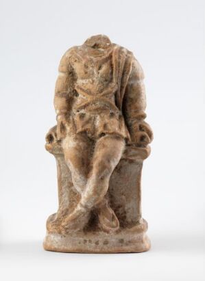  A small, weathered statue of an unidentified seated figure with crossed legs and hands holding an unidentifiable object. The statue is carved from a light tan or beige material with a rough texture, suggestive of stone or ceramic, and is set against a plain off-white background. Artist name and title are unknown.