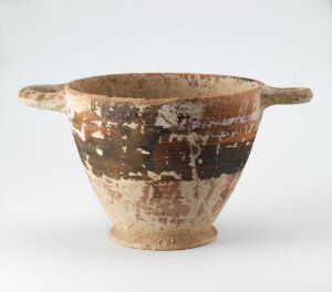  An ancient ceramic bowl with a cream base and horizontal bands of dark brown and black, featuring two small handles, on a plain background. The bowl shows signs of wear and aging. Artistname and title are unknown.