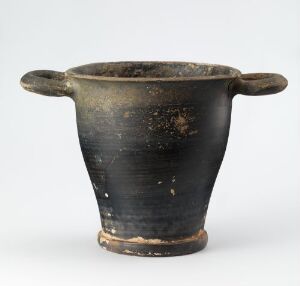  An ancient ceramic vessel with a black glaze finish and horizontal ridges, featuring two extended handles, showing signs of wear like chips and surface abrasions, against a neutral background.