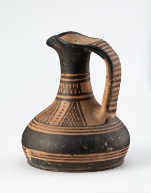  An ancient-style terracotta jug with a single handle and black geometric patterns on a light background. The jug has a flared opening, a wide body, and a narrow base, and is intricately decorated with parallel lines, triangles, and dots.