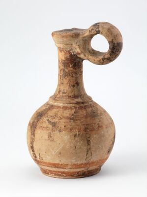  Ancient terracotta jug with a single handle, featuring a bulbous body and a narrow neck, against a neutral background. The jug has a pale, sandy hue with natural variations in color, including darker brown spots and streaks.