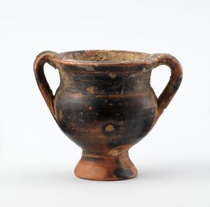  An ancient pottery vessel, possibly a cup or bowl, with a rounded body and two handles, exhibiting a dark, earthen tone with patches of terracotta or reddish-brown colour. The vessel rests against a plain white background, highlighting its silhouette and aged appearance.