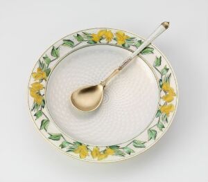  A decorative plate with a raised, woven-like texture at its center is adorned with a pattern of yellow flowers and green leaves around its rim, with a gold-toned spoon resting on top.