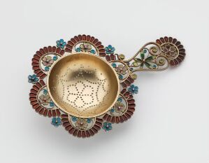  An ornate metal object, possibly historic, featuring a polished gold-colored center with lace-like patterns, surrounded by a halo of alternating deep red and pastel blue petal-shaped elements, with an ornamental handle-like extension showing matching hues.