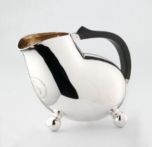  A modern silver pitcher with a sleek design, featuring a shiny metallic surface with a black matte handle and a thin gold band near the spout, supported by three spherical feet, against a light gray background. Artist name and title are unknown.