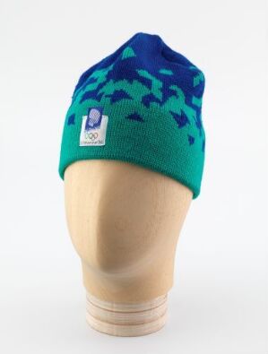  A bright green and deep blue beanie with a pixelated pattern transition is displayed on a featureless beige mannequin head against a white background. A white tag with purple and teal details is visible on the beanie’s rim.