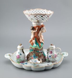  An ornate rococo-style ceramic centerpiece with a central tree-like structure supporting a perforated bowl, flanked by four small floral painted teapot-shaped containers on a tray with wavy edges and shell-like features.