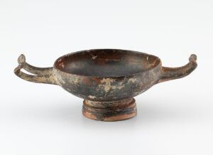  An image of an ancient bowl with looped handles on a circular base, displaying a dark brown patina with patches of lighter rust-like coloration and some greenish areas, indicative of age and possible metal oxidation.