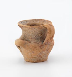  A small, textured clay pot with an uneven opening and a broken handle, displaying an earthy beige color with natural variations and speckles, suggesting an ancient or handcrafted origin.