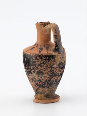  An ancient terracotta pottery jug with a rounded body, a single handle, and a flared opening. The jug has a mottled pattern of dark brown and orange splotches over a base of warm clay color, suggesting age and historical utility.