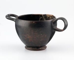  An ancient black ceramic cup with two handles and a worn terracotta interior, displayed against a white background.