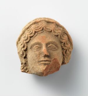  A fragmentary ancient terracotta sculpture depicting a face with stylized wavy hair and remnants of red pigment on the right cheek and lips, set against a light grey background.