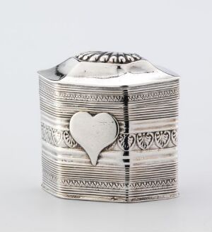  A silver hexagonal-shaped box with intricate engravings and a raised heart on one side, set against a plain light grey background.