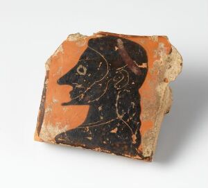  Fragment of an ancient terracotta object, likely a pottery shard, showing a stylized black figure in profile facing left on a reddish-orange background, with details indicating facial features and an earring.