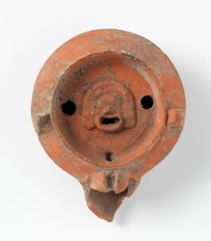  An ancient terracotta pottery artifact with a stylized face in the center, including round eye cavities, a raised nose, and a subtle mouth. The piece is a warm earthy red with hints of orange and has a rough texture that suggests historical significance.