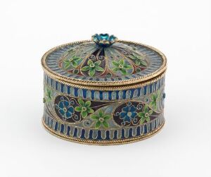  A decorative circular box with a rich cobalt blue and gold color scheme, featuring intricate patterns and floral designs in enamel, with a domed lid topped by a floral bud-like ornament.