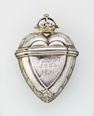  A metallic silver heart-shaped object, possibly a locket, with an engraved Cyrillic inscription on a banner across its center, topped with a decorative crown element, against a light gray background.