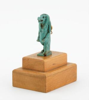  A turquoise statuette of the Egyptian goddess Taweret, depicted with a hippopotamus head, lion limbs, and crocodile tail on a light brown, stepped wooden pedestal. The artist's name and title of the piece are unknown.