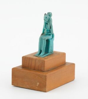  A small, turquoise-blue faience amulet of the Egyptian goddess Sekhmet, with a lioness's head on a woman's body, seated on a throne, displayed on a light brown wooden base. The title of the piece is "Amulett med gudinnen Sekhmet," and the artist is unknown.