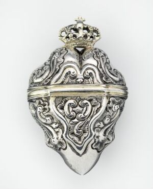  An ornate, embossed silver heart with a small crown on top, featuring a symmetrical, scroll-like pattern and a horizontal band in the middle against a plain white background. Artist name and title are unknown.