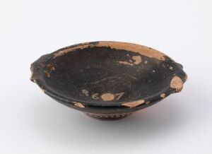  An ancient ceramic bowl with a black glaze and terracotta patches on a white background. The bowl has a wide, shallow shape with a cogwheel-like rim and is visibly aged with chips and wear marks.