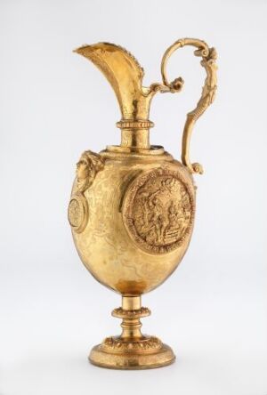  An ornate golden pitcher with classical embossed medallions, featuring a curved handle and an elaborate leaf-shaped spout, displayed against a neutral background. Artist name and title are unknown.