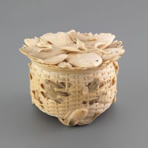  An exquisitely carved, pale-toned ornamental basket with an open top, featuring an interlocking lattice pattern and adorned with overlapping leaf-like carvings around the rim, set against a solid gray background.