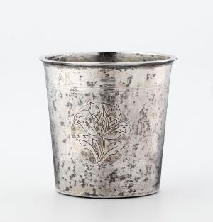  A tarnished silver-colored metal cup with an engraved symmetrical floral design on the front, exhibiting aged patina and an antique appearance against a neutral background. Artist name and title are unknown.