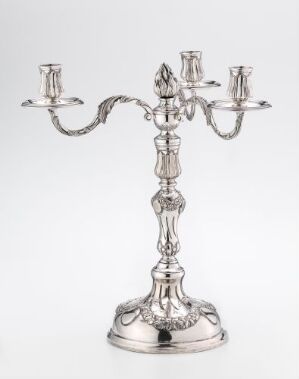  A silver three-branched candelabra with intricate designs, featuring a raised central candle holder topped with a flame-shaped finial, and two symmetrical side candle holders on elegant, curving arms, against a plain light background.