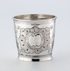  An intricately engraved silver cup with a stylized empty cartouche at the center, surrounded by ornate scroll and plant motifs, on a neutral background.