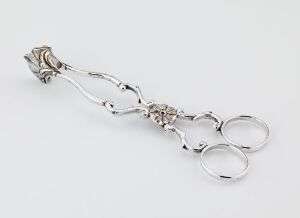  A polished silver sugar tong, titled "Sukkerklype" by Andreas Ferdinand Schumann, features an ornately embossed central detail with curved arms and rounded clamping sections, elegantly designed and resting on a neutral background.