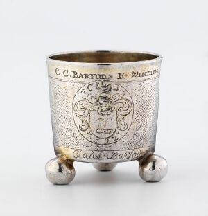  A Simon Mathiesen-designed silver beaker titled "Beger" with three spherical feet, showcasing detailed engraving including a central coat of arms, surrounded by intricate stippled patterns, and featuring engraved lettering, conveying a sense of historic craftsmanship and elegance.