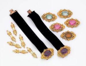  A collection of vintage-style jewelry featuring two black velvet chokers with gold-tone ends and various round brooches with colorful central stones surrounded by intricate gold-tone frames, along with three pairs of matching gold-tone earrings or pins, all displayed against a light background.