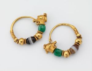  A pair of ancient-style circular gold earrings with stylized lion's head clasps and adorned with green and brown beads, against a pale neutral background.