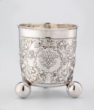  A silver ornamental cup with intricate embossing and a monogram in the center, supported by three spherical feet, set against a neutral light-colored background.