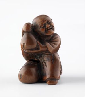  A small, chocolate-brown carved wooden figurine of a person seated with their head resting on their knees and arms wrapped around, set against an off-white background. The figure exhibits a polished finish and shows a detailed expression of contemplation or distress.