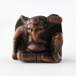  A small, dark brown intricately carved sculpture of a figure hugging its knees, with detailed hair and facial features, set against a light background. Artist name and title are unknown.