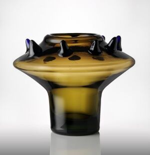  A glass vase with a flared, wave-like rim accentuated by dark blue or purple pointed tips, featuring a color palette that transitions from opaque black at the base to a transparent amber in the center, with swirls of brown and black, and set against a gradient background fading from white to dark. Artist name and title unknown.