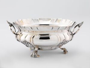  An oval-shaped silver sugar bowl titled "Sukkerskål" by Niels Rasmussen Holst, featuring chiseled designs and ornate handles, resting on three intricate feet. The polished silver has a reflective, high shine finish.