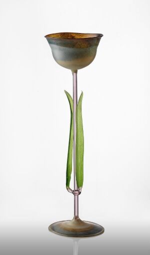  An artistic glass goblet with a golden amber to burnt orange bowl, slender green leaf-like stem, and a muted gold base against a plain white background. The artist's name and title are unknown.