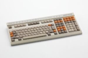  A vintage Norsk Data "ND NOTIS" keyboard with a light grey metallic base and keys, featuring distinctive orange function keys, set against a simple light background.