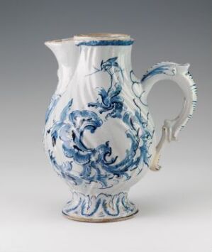  A white-glazed faience pitcher with intricate hand-painted cobalt blue decoration, crafted by Herrebøe Fajansefabrikk. The pitcher has an ornate handle and a voluptuous body adorned with swirl-like patterns suggesting stylized foliage or floral motifs.