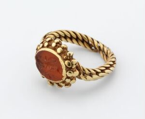  A gold ring with a twisted rope-like band and beaded details features an oval carnelian stone set at the top with an engraved design.