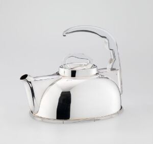 This is a photograph of "Strykejernet," a sterling silver teapot with a perspex detail designed by J. Tostrup. The teapot is set against a neutral grey background and reflects a high sheen, illustrating the sterling silver material and the artistry of the smiing technique. The piece demonstrates modern design elements with functional intent within the realm of design/kunsthåndverk.