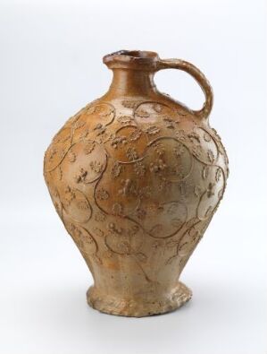  An ornate amber-colored glass jug with intricate engravings, featuring a single handle and a narrowing neck, set against a neutral off-white background. Artist name and title of the work are unknown.