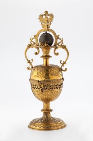  A ceremonial golden goblet with intricate engravings and decorative handles, featuring a contrasting dark sphere at the top, against a simple white background. Artist name and title are unknown.