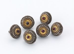  A collection of seven antique-looking buttons with a central purplish-red gemstone, surrounded by gold rings and white seed pearls, against a light gray background.