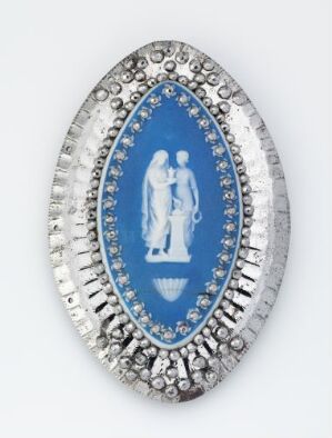  An oval jewelry piece with a deep blue center engraved with a white silhouette of a classical man and woman, surrounded by a sparkle of clear gemstones set in a silver or white gold frame.