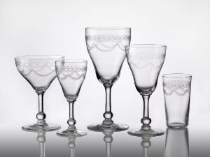  A set of five transparent glass tableware pieces with an intricate etched design on a gradient background, including a champagne coupe, two wine glasses of different heights, a wide-brimmed goblet, and a small tumbler, all on a reflective surface. Artist name: Ukjent. Title: Unknown.