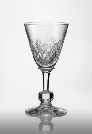  A delicately crafted clear glass goblet featuring skjærslipt and fasettslipt designs. The goblet has a wide top, tapering to a short stem with a spherical center, and a flat round base, displayed against a gradient background of white to grey.